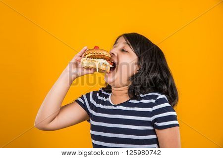 cute indian girl eating burger, small asian girl and burger, isolated over yellow background