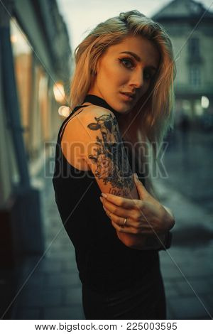 Young woman portrait with tattoo on shoulder standing on city street in evening. In background there are city lights.