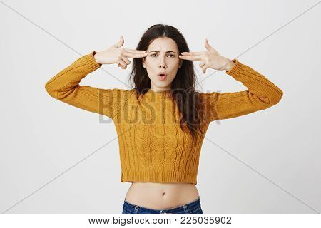 Annoyed and tired cute european female showing gun signs near temples folding open mouth and frowning, standing over gray background in trendy cropped sweater. Girl can not bear this pressure anymore.