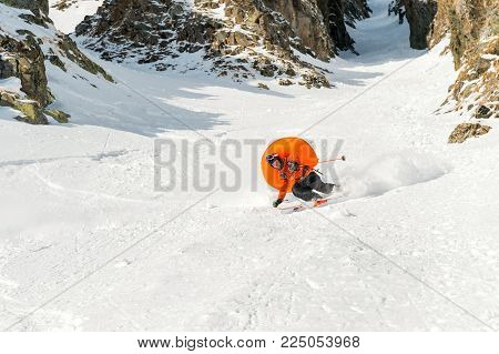 A male skier freerider with a beard descends the backcountry at high speed from the slope leaving a trail of snow powder with the opened anti-avalanche pillow ABS behind him against the background of epic rocks. The concept of freeriding culture and backc