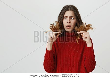 Horizontal portrait of young amazed stunned female model wearing red sweater looking with puzzlement and confusion at her dark long hair, being dissatisfied with work of her hairdresser. Negative emotions and reaction