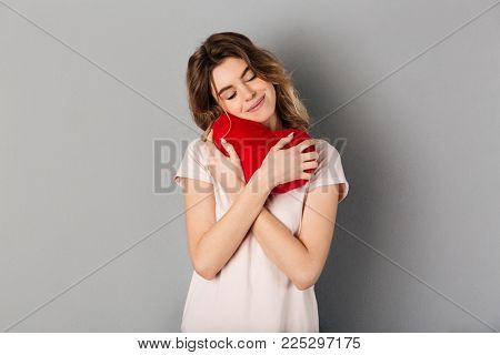 Image of Pleased woman in t-shirt holding joy heart with closed eyes over grey background