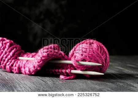 closeup of a pink knit cap in knitting progress, next to a ball of yarn and a pair of  needles, on a rustic wooden table against a dark background with some blank space on top