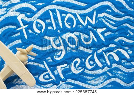 Follow Your Dream Inscription On A Blue Colored Sand With Waves And Wooden Plane, Top View, Flat Lay