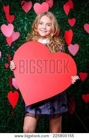 Happy pre-teen girl holding red heart surrounded by little hearts over lawny background. First love. Valentine's Day. 