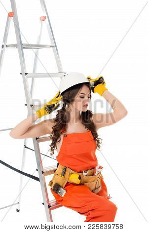 Brunette Workwoman In Overalls And Safety Helmet Posing Near Ladder, Isolated On White