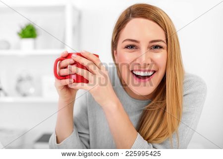 A Cute Young Woman Is Holding A Red Cup Sitting At A Table In A White Office. Coffee Break - Smiling