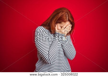 Young redhead woman wearing strapes navy shirt standing over red isolated background with sad expression covering face with hands while crying. Depression concept.
