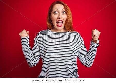 Young redhead woman wearing strapes navy shirt standing over red isolated background celebrating surprised and amazed for success with arms raised and open eyes. Winner concept.