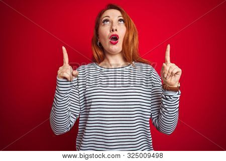 Young redhead woman wearing strapes navy shirt standing over red isolated background amazed and surprised looking up and pointing with fingers and raised arms.