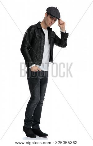 side view of an attractive casual man wearing a black leather jacket and hat standing with one hand in pocket and fixing his hat while leaning forward confident against white studio background