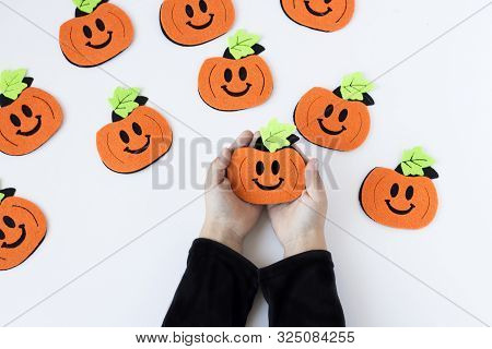 Top View Of Halloween Crafts, Felt Handmade Pumpkins On White Paper Background In The Hands Of A Chi