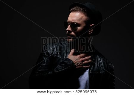 dramatic cool guy wearing leather, sunglasses and hat, holding hands on neck and looking to side, on grey background in studio