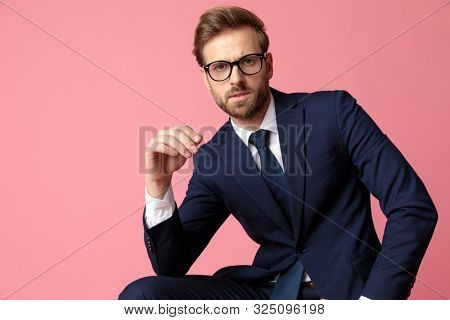 elegant formal business man in navy suit and glasses is sitting and resting his hands on his laps while  looking at camera serious on pink studio background