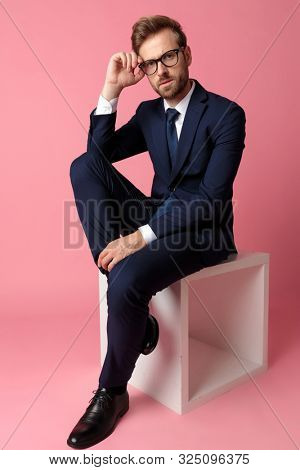 handsome formal business man in navy suit  sitting and resting  hands on laps and one leg resting on chair while fixing glasses serious on pink studio background