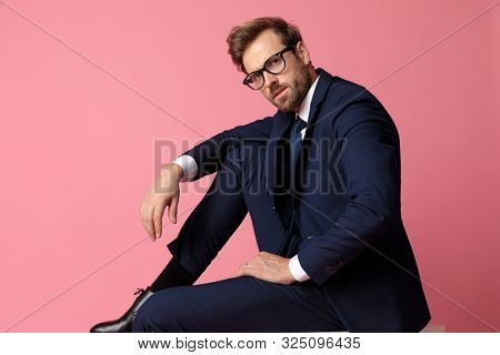 side view of a handsome formal business man in navy suit with eyeglasses  sitting with one leg resting on the chair and hands resting on his laps while looking at camera  on pink studio background