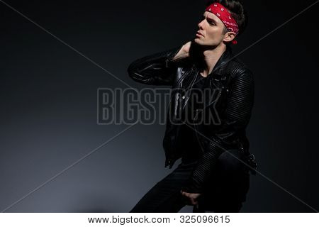 cool fashion model wearing leather jacket and red bandana, looking to side and holding hand on neck, sitting on grey background in studio