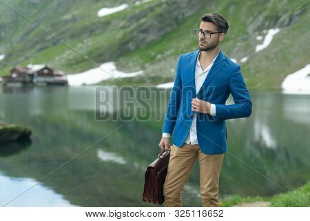 smart casual man wearing adjusting coat and holding suitcase, looking confused to side, outdoor in nature at Balea Lake, Romania