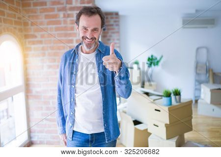 Middle age senior man moving to a new house packing cardboard boxes doing happy thumbs up gesture with hand. Approving expression looking at the camera with showing success.