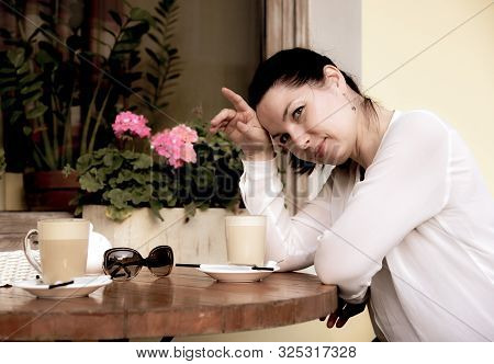 A Beautiful Woman Sits At A Table In A Street Cafe And Looks Slyly At The Camera. Two Cups Of Coffee