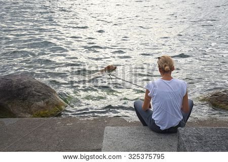 Zug, Switzerland 29.09.2019: A Sitting Blonde Woman In White Shirt And Jeans On The Lakeshore Boardw