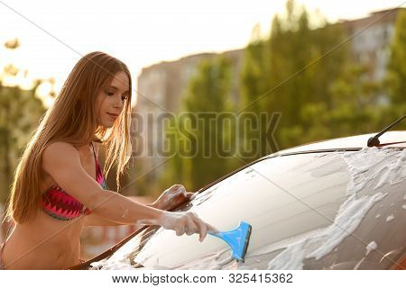 Young Woman In Swimsuit Washing Windshield Of Car With Squeegee Outdoors