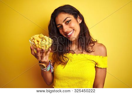 Young beautiful woman holding bowl with dry macaroni pasta over isolated yellow background with a happy face standing and smiling with a confident smile showing teeth