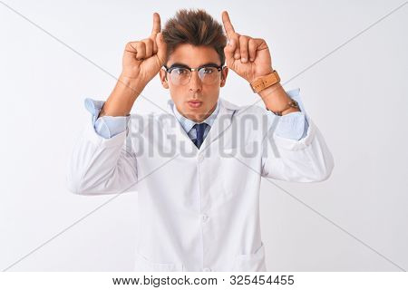 Young handsome sciencist man wearing glasses and coat over isolated white background doing funny gesture with finger over head as bull horns