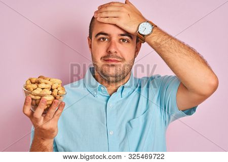 Young man holding bowl with peanuts standing over isolated pink background stressed with hand on head, shocked with shame and surprise face, angry and frustrated. Fear and upset for mistake.