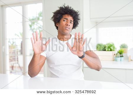Young african american man wearing casual white t-shirt sitting at home Moving away hands palms showing refusal and denial with afraid and disgusting expression. Stop and forbidden.