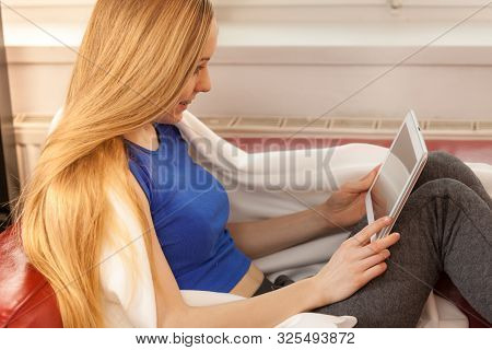 Young Woman Lying On Sofa Under Warm Blanket, Relaxing Using Tablet, Reading News And Checking Socia
