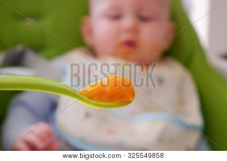 Spoon Full Of Carrot Pure And A Blurry Baby On Background. Selective Focus On The Spoon. Baby First