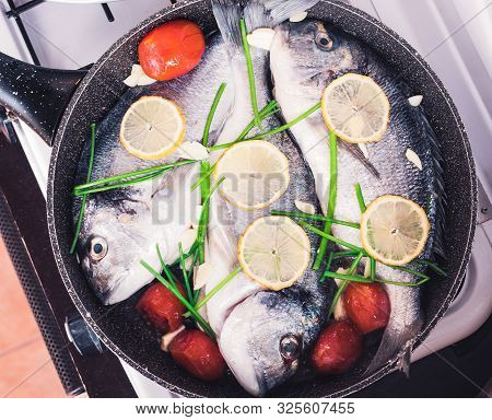 Raw Orata Three Whole Fish On Frying Pan Cooking With Fresh Lemon Slices, Tomatoes And Greenery Ital