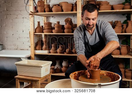 Handsome Young Potter Man In A Gray Sweater And A Black Apron Sculpts Clay Pot On A Potters Wheel In