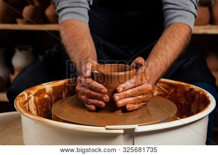 Handsome Young Potter Man In A Gray Sweater And A Black Apron Sculpts Clay Pot On A Potters Wheel In
