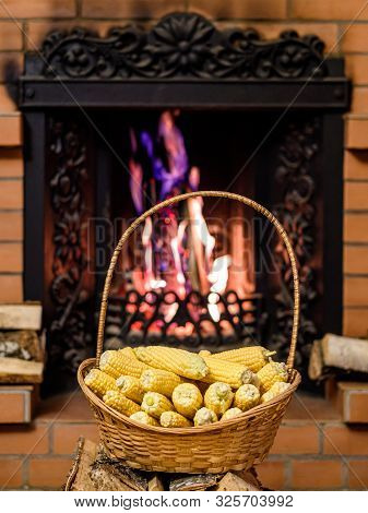 A Wicker Basket To The Top Filled With Ears Of Ripe Corn. In The Background Firewood Burning In The