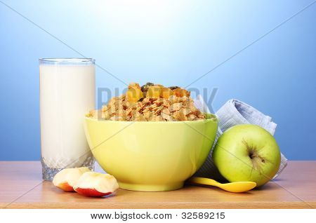 leckere Cornflakes in grüner Schale, Äpfel und Glas Milch auf Holztisch auf blauem Hintergrund