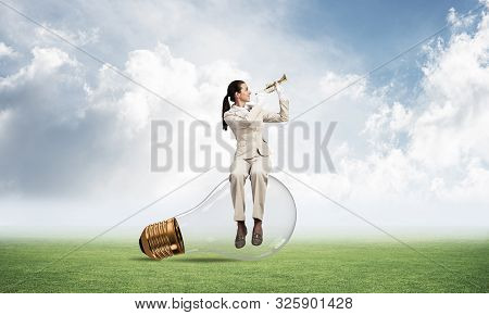 Woman Sitting On Big Light Bulb And Playing Trumpet Brass. Young Lady In White Business Suit And Glo