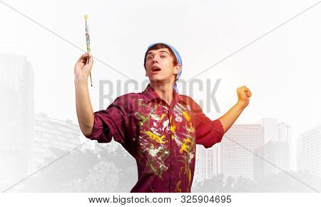 Young Emotional Artist Gesturing With Paintbrush. Happy Painter In Shirt And Bandana Standing On Bac