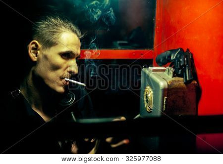 A Man Of Criminal Appearance In A Payphone Booth At Night. Talking On The Phone With A Revolver And