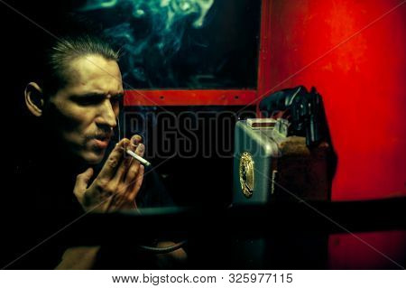 A Man Of Criminal Appearance In A Payphone Booth At Night. Talking On The Phone With A Revolver And
