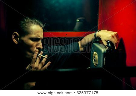 A Man Of Criminal Appearance In A Payphone Booth At Night. Talking On The Phone With A Revolver And