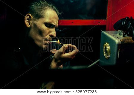 A Man Of Criminal Appearance In A Payphone Booth At Night. Talking On The Phone With A Revolver And