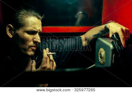A Man Of Criminal Appearance In A Payphone Booth At Night. Talking On The Phone With A Revolver And