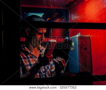 A Man Of Criminal Appearance In A Payphone Booth At Night. Talking On The Phone With A Revolver And