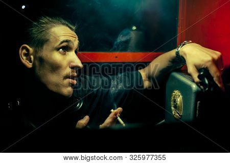 A Man Of Criminal Appearance In A Payphone Booth At Night. Talking On The Phone With A Revolver And