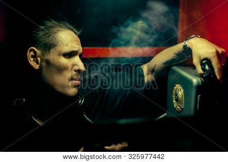 A Man Of Criminal Appearance In A Payphone Booth At Night. Talking On The Phone With A Revolver And