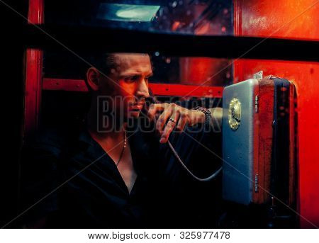 A Man Of Criminal Appearance In A Payphone Booth At Night. Talking On The Phone With A Revolver And
