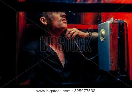 A Man Of Criminal Appearance In A Payphone Booth At Night. Talking On The Phone With A Revolver And