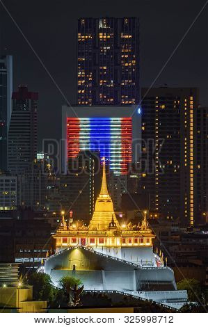 Golden Mount Or Wat Saket, Phukhao Thong, And Skyscraper Buildings With Thailand Flag At Night In Ba